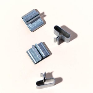 May include: Four small, silver-colored metal clips are arranged on a light beige surface. Two clips are rectangular with a folded design, while the other two have a U-shaped design with a small tab extending from each end. The clips are made of a shiny, metallic material.