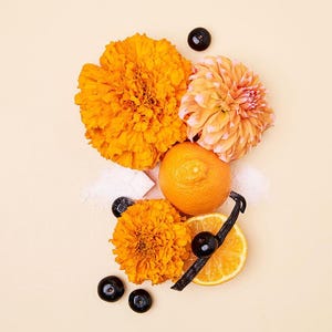 May include: An overhead shot features vibrant orange flowers, a whole orange, and a sliced orange with a vanilla bean. Dark berries and sugar cubes are scattered around the arrangement on a light beige background.