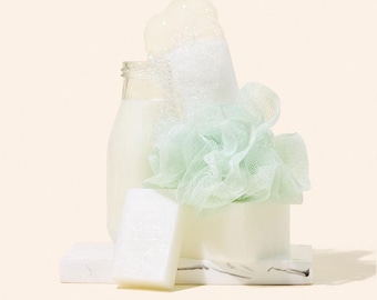 May include: A still life composition featuring bath products. A clear glass bottle filled with a milky liquid is next to a white bar of soap and a light green mesh loofah. Bubbles cascade from the bottle.