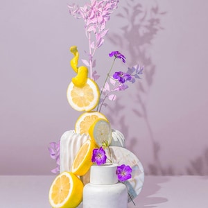 May include: A still life composition featuring sliced lemons, purple flowers, and pink foliage arranged on white ceramic vases and a marble-patterned disc. The lemons are bright yellow, contrasting with the purple and pink elements. The background is a soft lavender color.