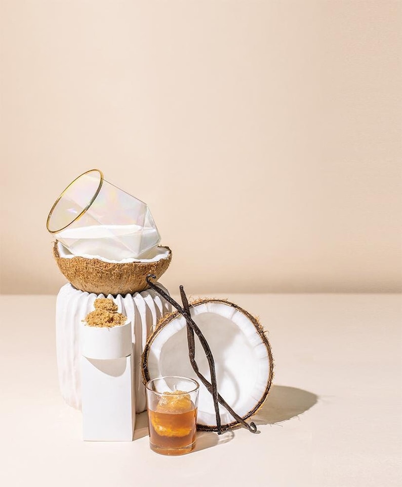 May include: A still life arrangement featuring a halved coconut, a geometric glass with a gold rim, and a small glass of amber liquid. Also present are vanilla beans and a white, textured vase. The scene is set against a neutral background.