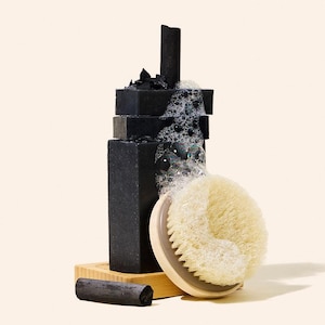 May include: A stack of black charcoal soap bars with bubbles, a wooden brush, and a piece of charcoal. The soap bars are on a small wooden stand. The brush has a light wood handle and light-colored bristles.