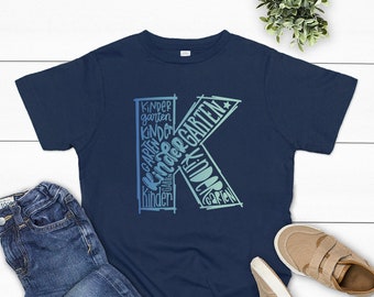 以下が含まれることがあります： 紺色のTシャツに、青と緑の大きなKの文字が、さまざまなフォントとサイズで「kindergarten」という言葉で構成されています。