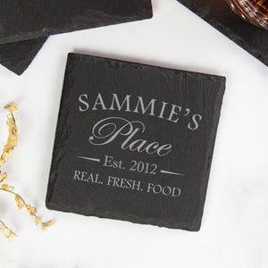 以下が含まれることがあります： 「SAMMIE'S Place Est. 2012 REAL. FRESH. FOOD」の文字が薄いグレーで書かれた、正方形の黒いスレートコースター。コースターは他のコースターと液体の入ったグラスに囲まれています。