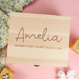 May include: A light-colored wooden box with the name "Amelia" in a cursive font. The box includes the date, time, weight (7 lbs 12 oz), and length (20 in). Two wooden blocks and a floral fabric are also visible.