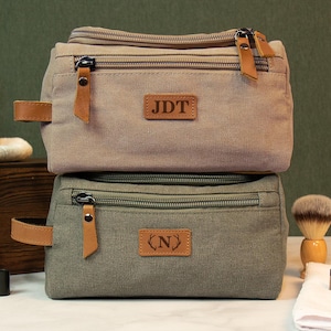 May include: Two canvas toiletry bags with leather accents. The top bag is light brown with the initials "JDT" on a leather tag. The bottom bag is dark green with the initial "N" on a leather tag.