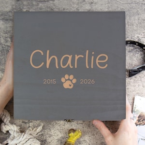 May include: A gray wooden pet keepsake box with the name "Charlie" in a light brown script. The box features a paw print and the years 2015 and 2026. A black dog collar and a photo are visible.