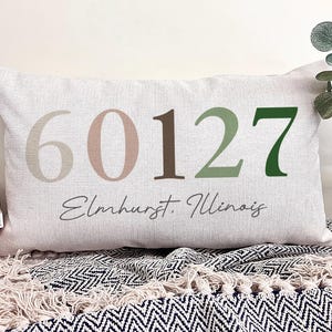 May include: Rectangular beige pillow with the numbers "60127" in shades of taupe, brown, and green. The text "Elmhurst, Illinois" is written below the numbers in a script font. The pillow is on a patterned blanket.