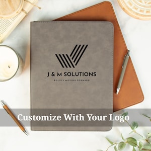 May include: A gray leather-like portfolio with the text "J & M SOLUTIONS" and a logo. The phrase "BOLDLY MOVING FORWARD" is below the text. The words "Customize With Your Logo" are at the bottom. A pen and a notebook are also visible.