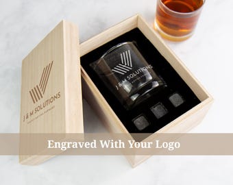Peut inclure: Coffret cadeau en bois avec un verre à whisky et des pierres à whisky. Le verre et le couvercle de la boîte portent le texte "J & M SOLUTIONS" et un logo. La boîte est ouverte, révélant un intérieur noir. Un verre de liquide ambré est en arrière-plan.