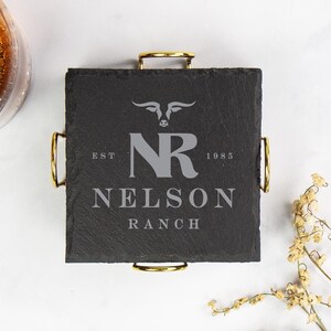 以下が含まれることがあります： 金色のハンドルが付いた正方形の黒いスレートコースター。テキスト「NELSON RANCH」とイニシャル「NR」と雄牛の頭のロゴが特徴です。コースターは飲み物用に設計されています。