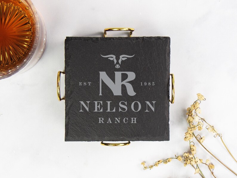 以下が含まれることがあります： 金色のハンドルが付いた正方形の黒いスレートコースター。テキスト「NELSON RANCH」とイニシャル「NR」と雄牛の頭のロゴが特徴です。コースターは飲み物用に設計されています。
