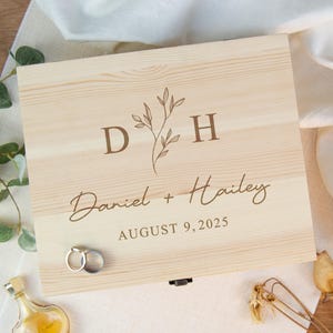 May include: A light-colored wooden box with engraved text and a floral design. The text reads "D H", "Daniel + Hailey", and "AUGUST 9, 2025". Two rings are on the box. A small bottle and greenery are in the background.