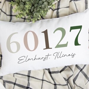 May include: White rectangular pillow with the numbers 60127 in various shades of brown and green. The word "Elmhurst, Illinois" is written in a script font below the numbers. The pillow is resting on a plaid blanket.