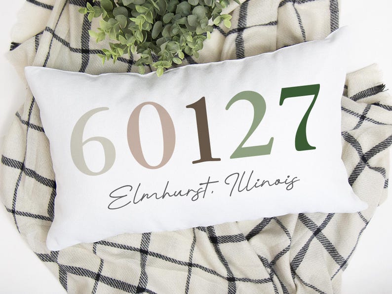 May include: White rectangular pillow with the numbers 60127 in various shades of brown and green. The word "Elmhurst, Illinois" is written in a script font below the numbers. The pillow is resting on a plaid blanket.