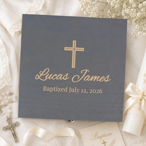 May include: A square gray wooden box featuring a gold cross, the name Lucas James, and the text Baptized July 12, 2026. Beside the box is a white candle with a gold cross, the text God Bless You, and a white ribbon, along with a silver rosary.