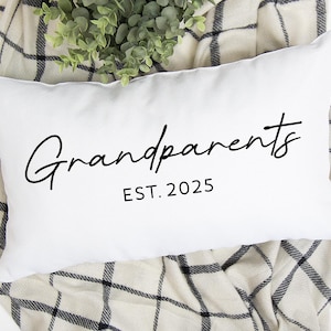 May include: White rectangular pillow with the word "Grandparents" in a cursive font and the text "EST. 2025" in a simple font. The pillow is on a plaid blanket with greenery.