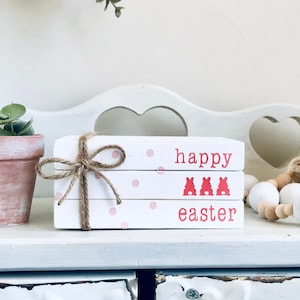 May include: A stack of three white books with pink polka dots and the words "happy easter" in red lettering. The books are tied together with twine and a bow. The books are sitting on a white shelf with a brown jar and a white and brown beaded garland.