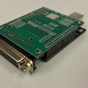 Könnte beinhalten: Eine grüne Platine mit einem schwarzen Stecker und einem silbernen 25-poligen Stecker. Die Platine hat einen USB-Anschluss und mehrere Reihen von Pins.