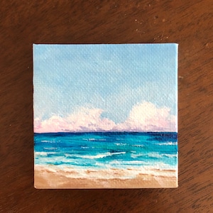 May include: A small painting of a beach scene with a blue sky, white clouds, and a turquoise ocean with white waves crashing on a sandy beach.