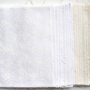 Könnte beinhalten: Ein Stapel weißer und beigefarbener Stoffquadrate mit einem dezenten Blumenmuster. Die Stoffquadrate sind in einem Farbverlauf von Weiß nach Beige angeordnet.