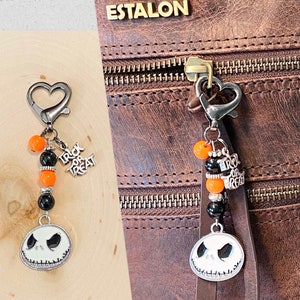 May include: A black and orange Halloween-themed keychain with a silver charm featuring a Jack Skellington-like face. The keychain has a silver heart-shaped clasp and is attached to a brown leather bag with the word "ESTALON" embossed on it.