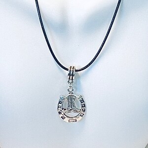 May include: A silver necklace with a black cord and a pendant shaped like a horseshoe with a boot inside. The pendant has a detailed design and is a great addition to any jewelry collection.