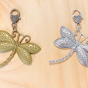 May include: Two dragonfly charms with intricate details. One is gold and the other is silver. Both have a heart-shaped clasp.