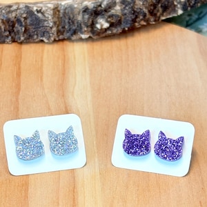 May include: Two pairs of cat-shaped stud earrings. One pair is silver glitter and the other is purple glitter.