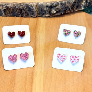 May include: Four pairs of heart-shaped earrings on white cards. The earrings are made of resin and have different designs, including glitter, pink hearts, and pink and green confetti.