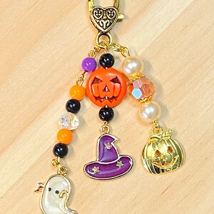 May include: A Halloween-themed charm with a gold-tone clasp and dangling charms. The charms include an orange jack-o'-lantern, a purple witch hat, a gold pumpkin, and a white ghost. The charm is adorned with beads in various colors.