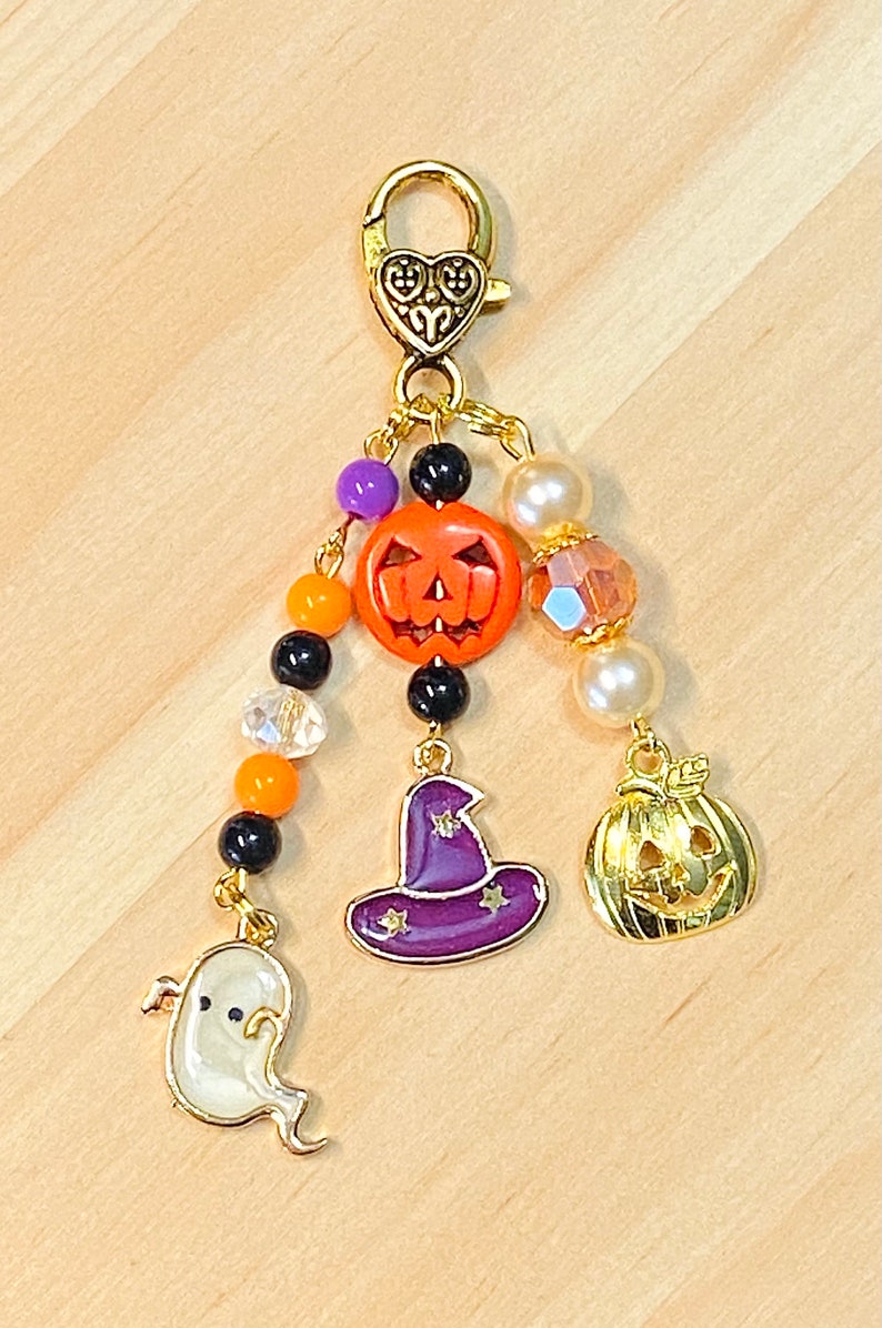 May include: A Halloween-themed charm with a gold-tone clasp and dangling charms. The charms include an orange jack-o'-lantern, a purple witch hat, a gold pumpkin, and a white ghost. The charm is adorned with beads in various colors.