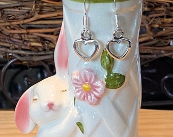 May include: A pair of silver heart-shaped earrings displayed on a white ceramic bunny figurine with pink ears and floral accents.