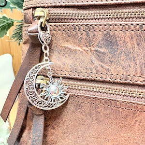 May include: A silver-toned metal charm featuring a crescent moon with intricate filigree cutouts and a sun emblem attached to the zipper pull of a brown leather bag. The charm hangs from a small heart-shaped connector and a short chain link.