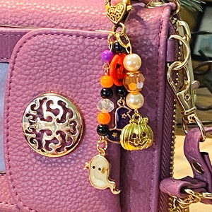 May include: A purple wallet with a gold-colored charm. The charm features Halloween-themed beads and charms, including a ghost, pumpkin, witch hat, and beads in orange, black, and white. The wallet has a gold-colored decorative emblem.