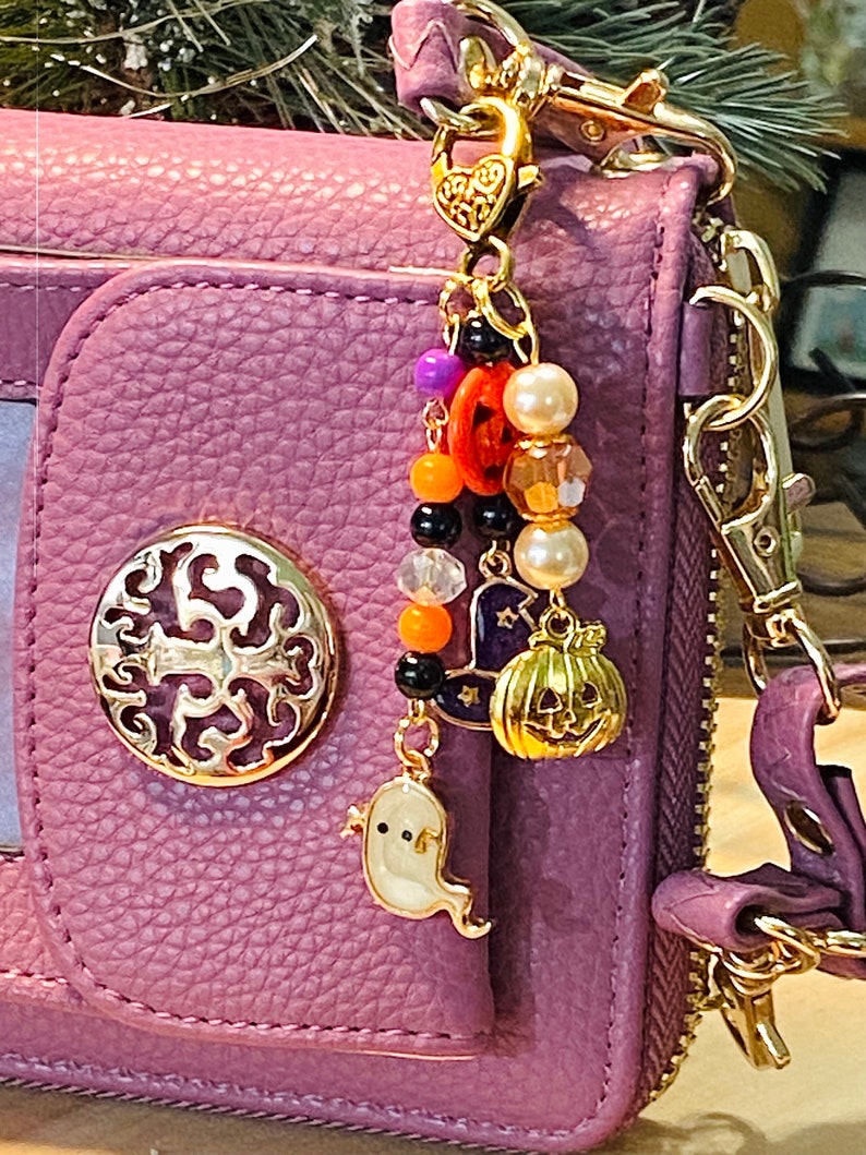 May include: A purple wallet with a gold-colored charm. The charm features Halloween-themed beads and charms, including a ghost, pumpkin, witch hat, and beads in orange, black, and white. The wallet has a gold-colored decorative emblem.