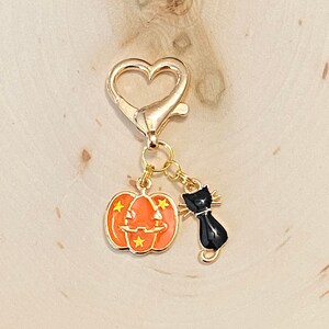 May include: A gold heart-shaped keychain with a black cat and orange pumpkin charm. The pumpkin has a jack-o'-lantern face and yellow stars.
