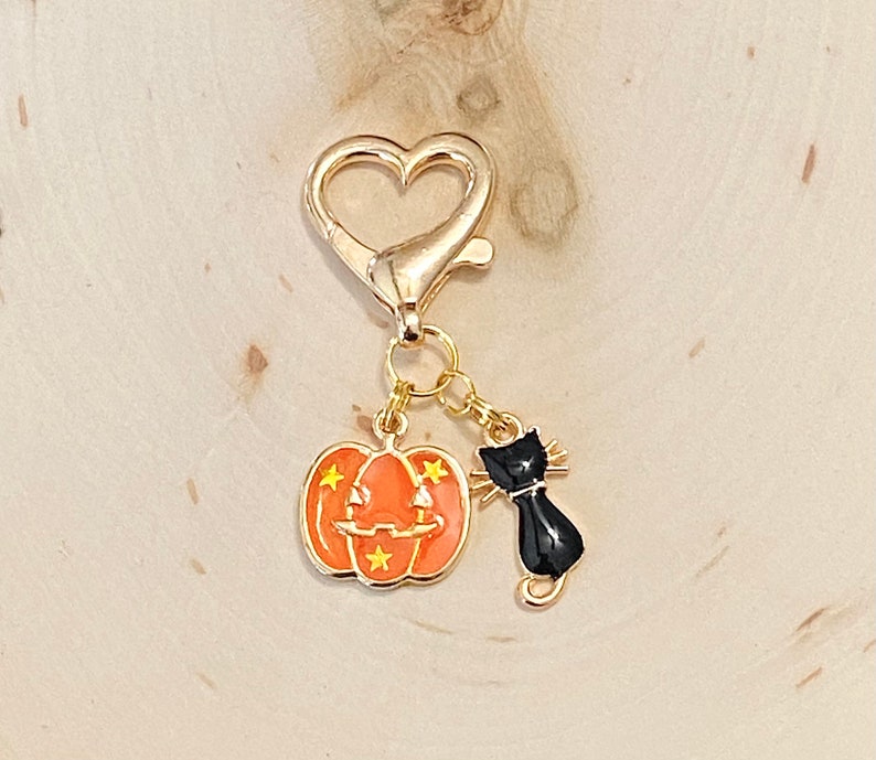 May include: A gold heart-shaped keychain with a black cat and orange pumpkin charm. The pumpkin has a jack-o'-lantern face and yellow stars.