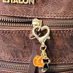 May include: A gold-toned zipper pull with a heart-shaped charm and a dangling Halloween-themed charm featuring a black cat and a pumpkin. The charm is attached to a brown leather bag.
