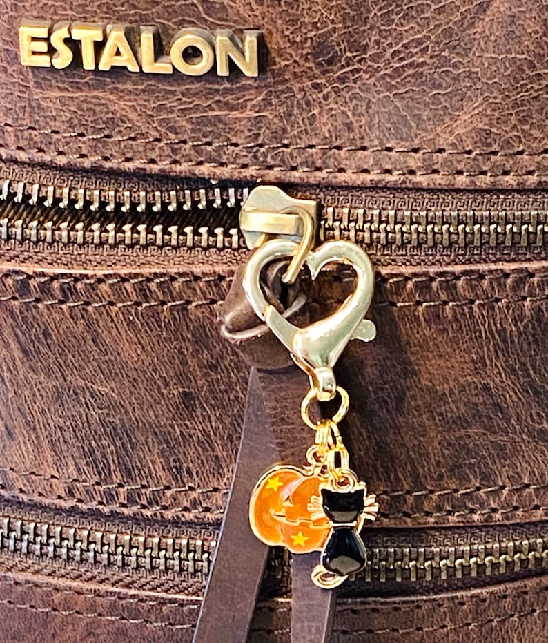 May include: A gold-toned zipper pull with a heart-shaped charm and a dangling Halloween-themed charm featuring a black cat and a pumpkin. The charm is attached to a brown leather bag.