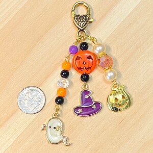 May include: A Halloween-themed charm with a gold-tone clasp and dangling charms. Charms include a smiling jack-o'-lantern, a witch's hat, a ghost, and a smaller gold pumpkin. The charm also features orange, black, purple, and clear beads.