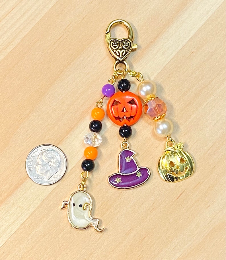May include: A Halloween-themed charm with a gold-tone clasp and dangling charms. Charms include a smiling jack-o'-lantern, a witch's hat, a ghost, and a smaller gold pumpkin. The charm also features orange, black, purple, and clear beads.
