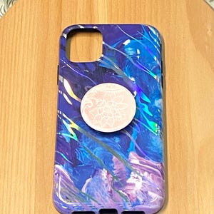 Puede incluir: Una funda de teléfono iridiscente de color morado, azul y blanco con un pop socket de diseño floral blanco.