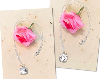 Könnte beinhalten: Zwei silberne Halsketten mit Anhängern. Ein Anhänger sagt "LOVE" und der andere hat ein Herzdesign. Beide Halsketten werden auf einer hellbraunen Oberfläche mit einer rosa Rose präsentiert.