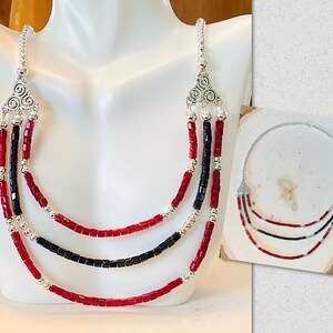 May include: A three-strand necklace with red and black beads and silver accents. The necklace has a silver pendant with a decorative design.