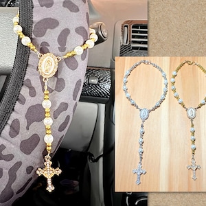 May include: A gold and pearl rosary hanging on a steering wheel. The rosary features a cross pendant, a center medal, and pearl beads. Two additional rosaries, one silver and one gold, are displayed on a wooden surface.
