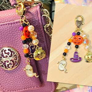 Op de afbeelding: Een goudkleurige Halloween-bedel met een karabijnsluiting. De bedel heeft een kralenketting, een lachende pompoen, een heksenhoed, een spook en een gouden pompoen. De bedel is bevestigd aan een paarse handtas.