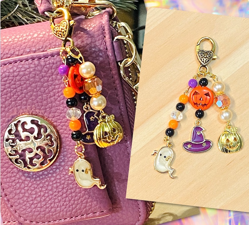 May include: A gold-toned Halloween-themed charm with a lobster clasp. The charm features a string of beads, a smiling jack-o'-lantern, a witch's hat, a ghost, and a golden pumpkin. The charm is attached to a purple purse.