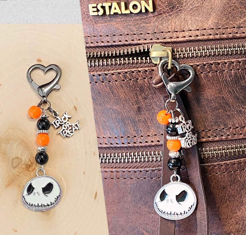 May include: A black and orange Halloween-themed keychain with a silver charm featuring a Jack Skellington-like face. The keychain has a silver heart-shaped clasp and is attached to a brown leather bag with the word "ESTALON" embossed on it.
