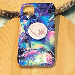 Puede incluir: Una funda de teléfono negra con un diseño abstracto de remolino colorido. Un corazón rosa con una huella de pata está en un soporte pop blanco unido a la funda.